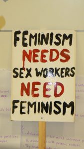A white sign on a stick that states: "Feminism NEEDS sex workers NEED feminism" , where both need/needs are in red.