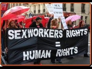 Protesters in France hold up signs that say "Sex Workers Unite!" and hold a large banner that say "SEXWORKERS RIGHTS = HUMAN RIGHTS".